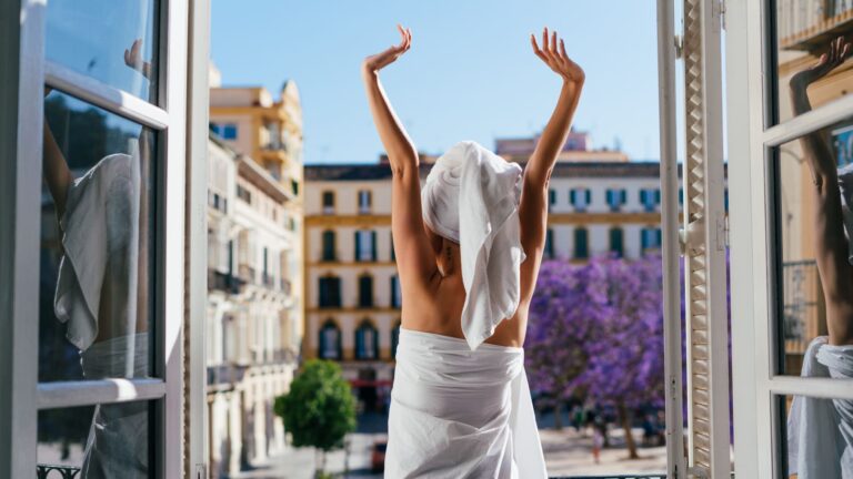A person stretches on an apartment balcony overlooking a European townscape in the morning light.