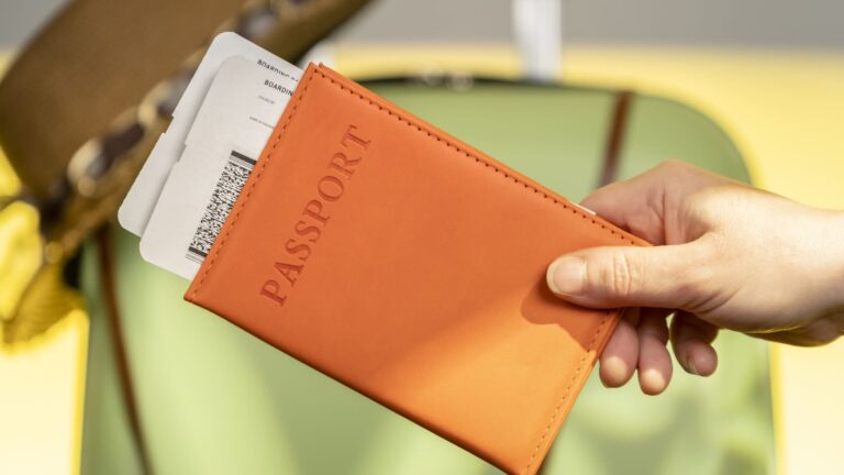 A traveler holds an orange passport wallet and a boarding pass next to a suitcase inside an airport terminal during the day.