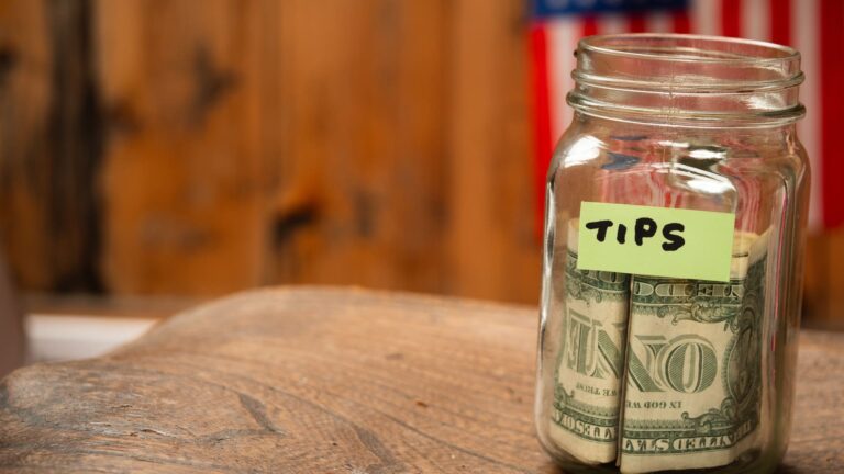 A tip jar filled with US dollar bills set against an American flag backdrop.