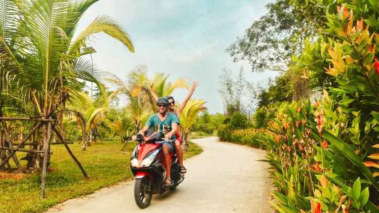 A smiling couple in helmets rides a motorbike scooter down a palm-lined road on Phu Quoc island, Vietnam.