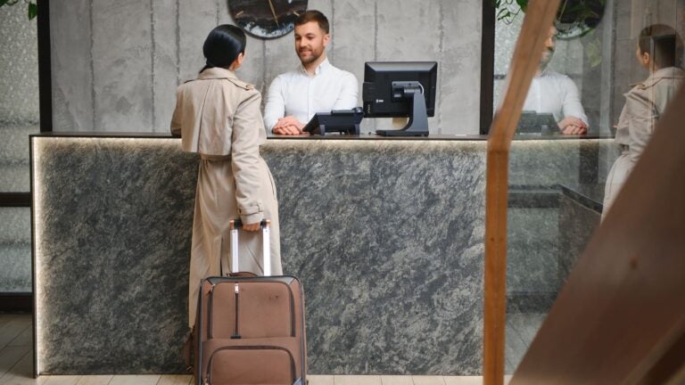 A woman with a rolling suitcase stands at a hotel front desk in the evening while a receptionist assists her.