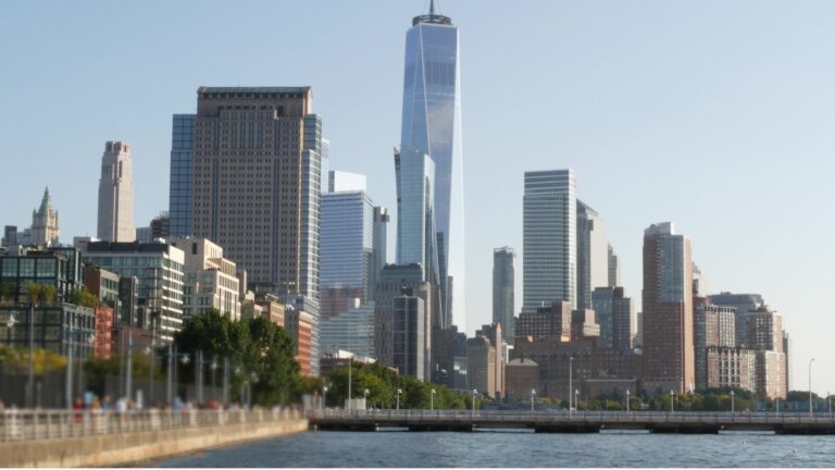 Lower Manhattan's Financial District skyline seen across the Hudson River, with people walking along the waterfront promenade.