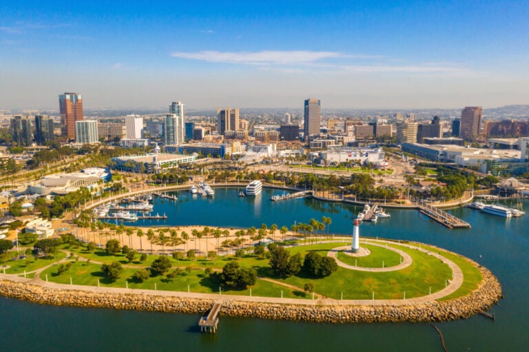 Aerial view of a curved waterfront with a white lighthouse, palm trees, and a marina of boats, with the Long Beach skyline behind it, Los Angeles, California.