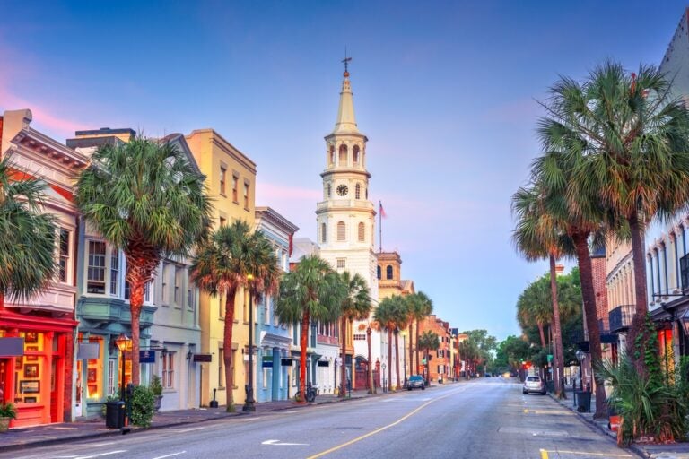 A quiet street in Charleston’s historic district, lined with palm trees and pastel buildings, with a white church steeple at the end of the road.
