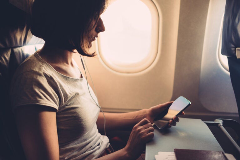 A traveler on a plane looking at a smartphone, with earbuds in, seated by the window.