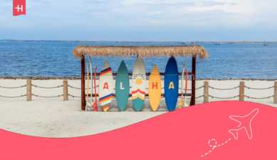Colorful surfboards spelling “Aloha” beneath a thatched shelter beside the ocean, with Holafly branding.