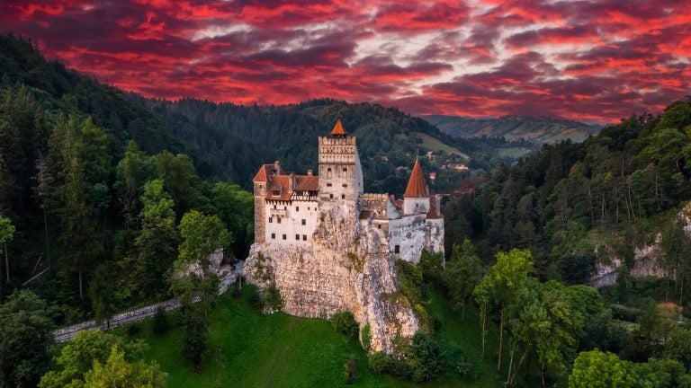 Bran Castle with its red-tiled towers and pale stone walls on a rocky hilltop, surrounded by forest and lit by the setting sun