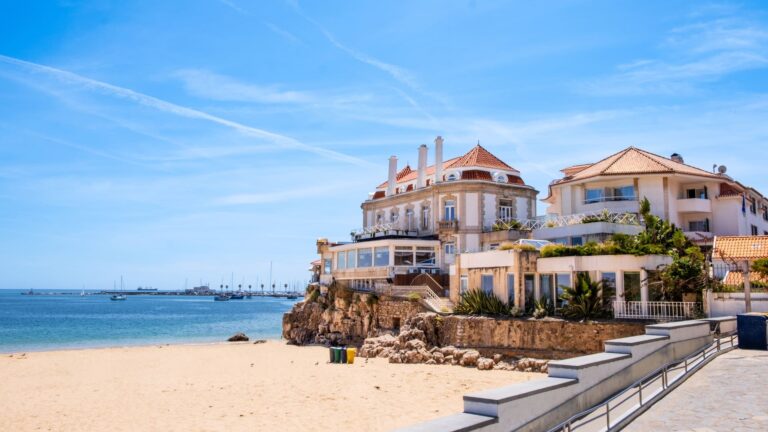 Small sandy cove at Praia da Rainha in Cascais, enclosed by rocky cliffs and white beachfront villas, with calm blue water under a clear sky.