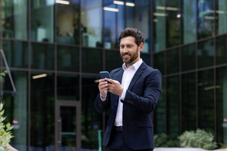 Man in a navy suit using a smartphone outside a glass office building.