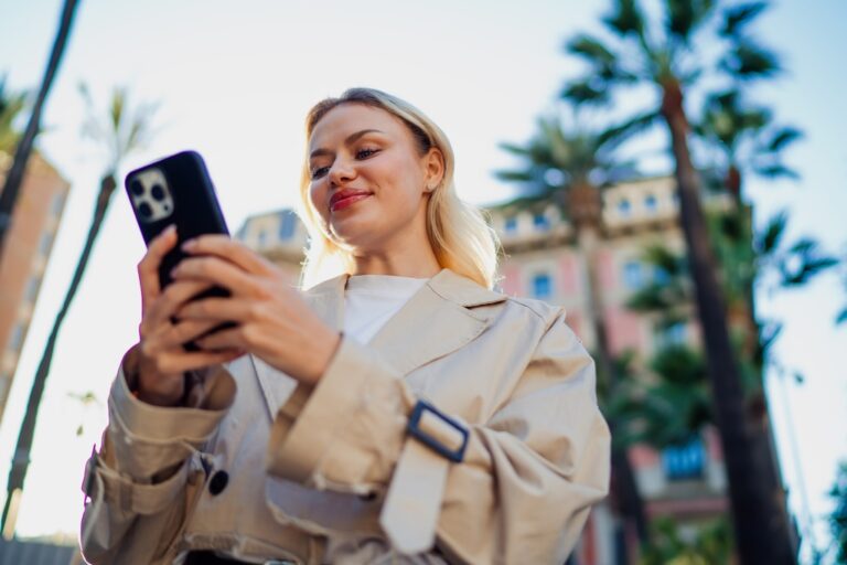 Woman in a beige coat using her smartphone outdoors with palm trees and buildings behind her.