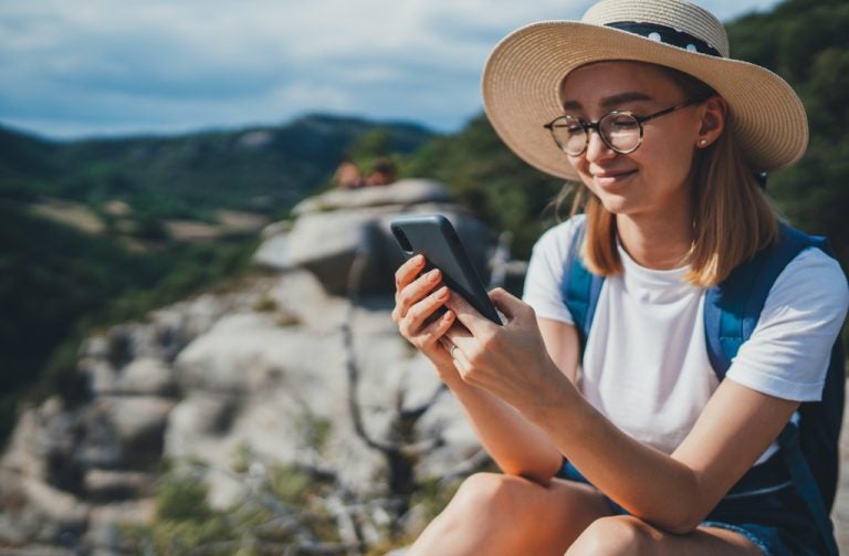 Traveler wearing a sun hat and backpack using her phone on a rocky hillside.