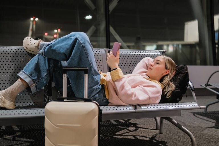 Traveler using her phone while reclining on airport seats beside a suitcase.