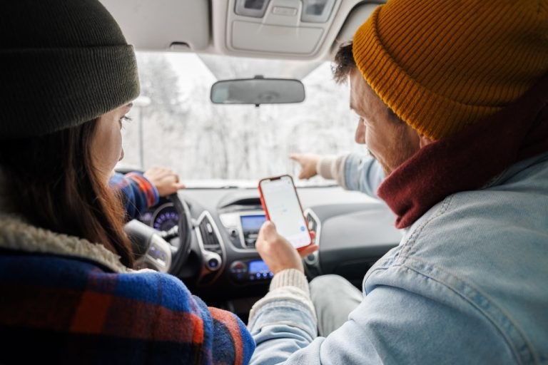 Passenger holding a smartphone and pointing ahead while a driver navigates a snowy road.