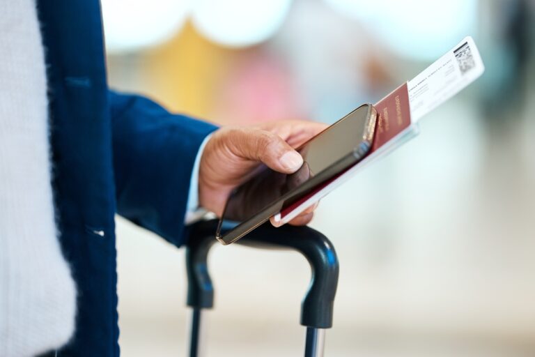 Traveler holding a smartphone, passport, and boarding pass above a suitcase handle.