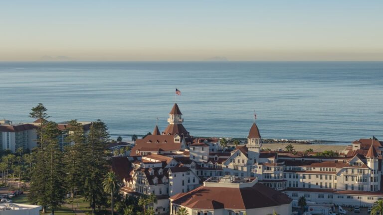 Drone shot of Hotel del Coronado, a sprawling Victorian resort with red turret roofs, beside the beach in San Diego.