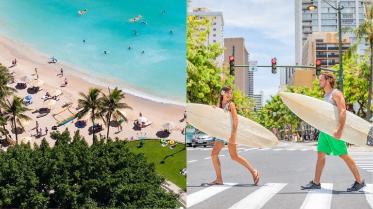 Aerial view of a palm-lined beach on Oahu and two surfers carrying boards across a city street.