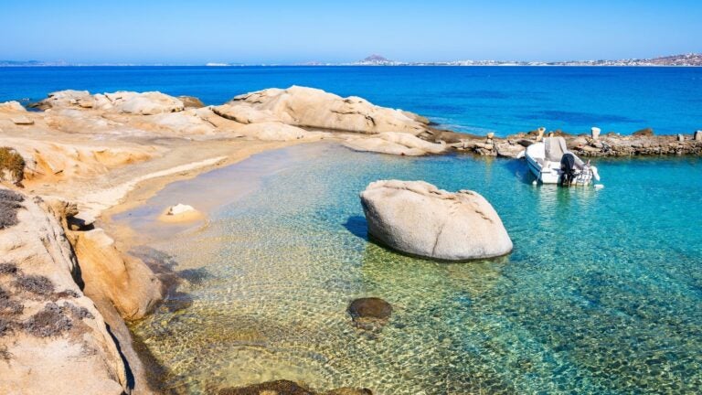 Smooth rocks scattered along the sandy shore of Orkos Bay on Naxos, with calm, clear turquoise water stretching out to sea