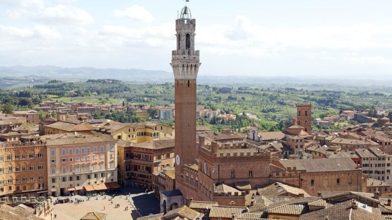 Aerial view of the shell-shaped Piazza del Campo and the tall Torre del Mangia bell tower, surrounded by terracotta rooftops in Siena, Italy.