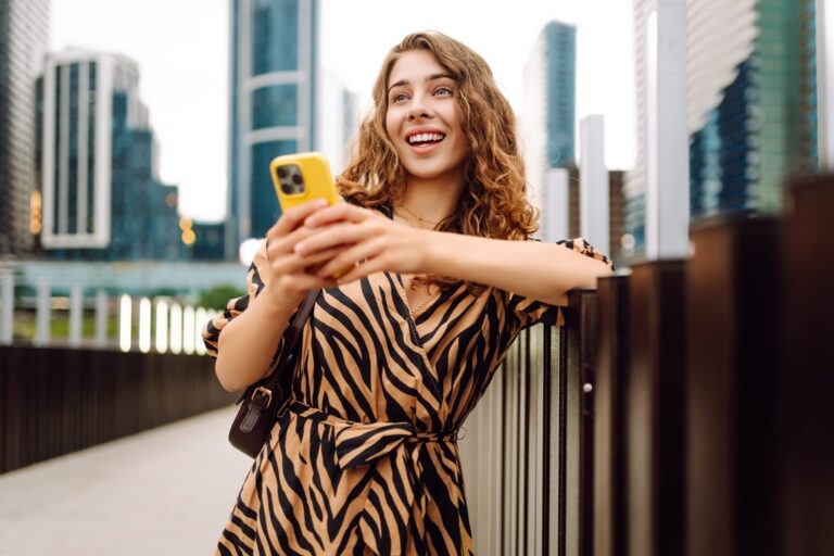 Smiling woman holding a yellow smartphone with city skyscrapers behind her.