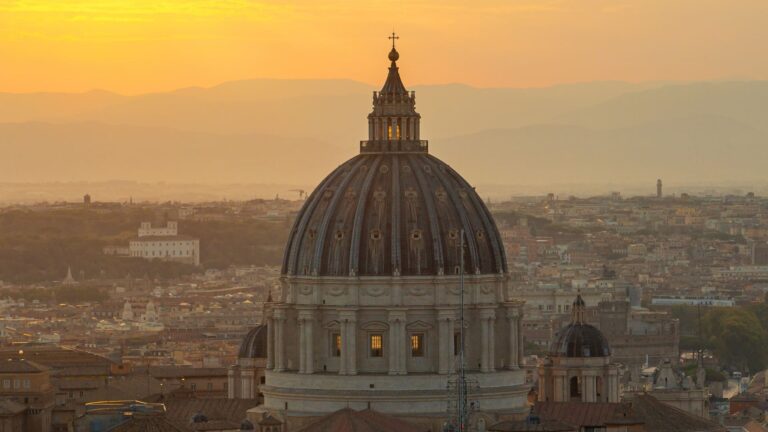 The dome of St. Peter’s Basilica rising above Rome’s rooftops at sunrise, with distant mountains on the horizon.