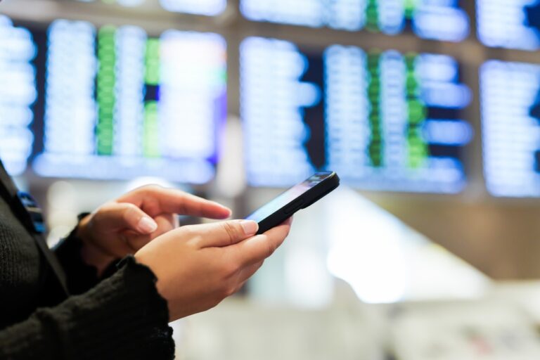 Close-up of a traveler using a smartphone in front of airport information screens.