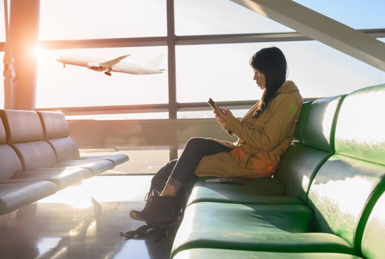 Woman using her smartphone on a green airport bench as a plane takes off outside.