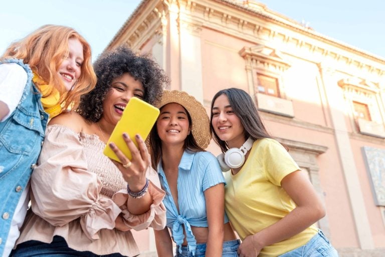 Four smiling young women outdoors in front of a pink historic building, gathered around a bright yellow smartphone that one of them is holding up.