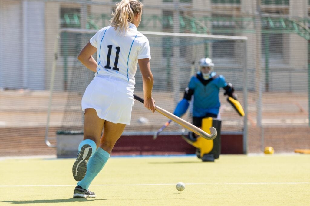 A player in white prepares to shoot at the goal during a field hockey match on a sunny day.