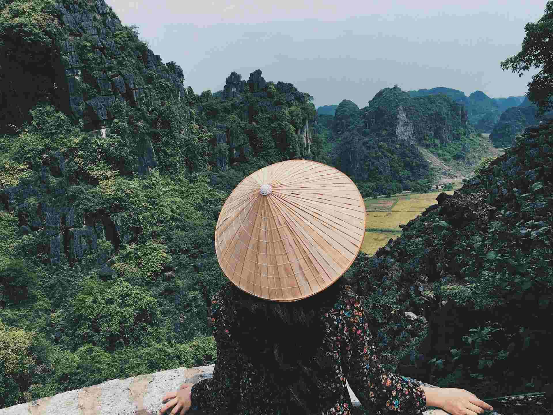 Eine Frau mit einem typisch vietnamesischen Hut sitzt vor einer mit Wald bedeckten Montagelandschaft.