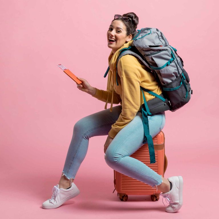 Eine lächelnde junge Frau sitzt auf ihrem Koffer, hat einen Rucksack auf dem Rücken und hält ihr Handy in der Hand.