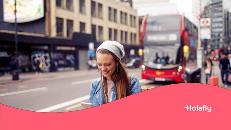 Eine junge Frau mit einem Bonet schaut in London auf ihr Handy, im Hintergrund ein roter Doppeldeckerbus.