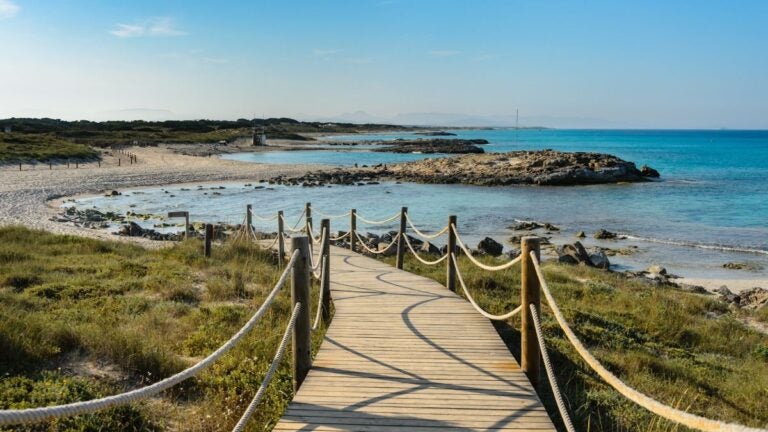 Holzsteg an einem ruhigen Strand auf Formentera mit türkisblauem Wasser und felsiger Küste.