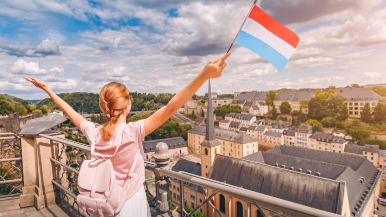 Eine glückliche Reisende hält die Flagge von Luxemburg und bewundert den Stadtteil Grund von einer Aussichtsplattform aus