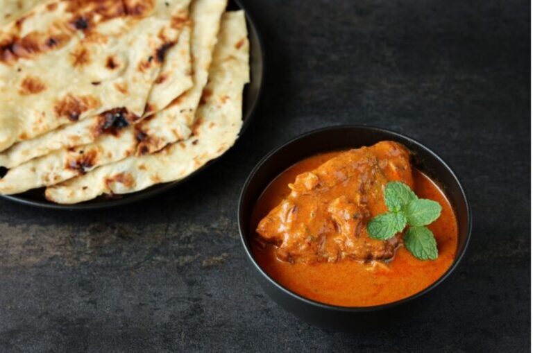 Eine dunkle Schale Butter Chicken, daneben gefaltetes Naan-Brot.