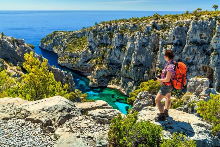 Eine Wanderin blickt von einer hohen Klippe hinab auf die Calanque d’En-Vau.