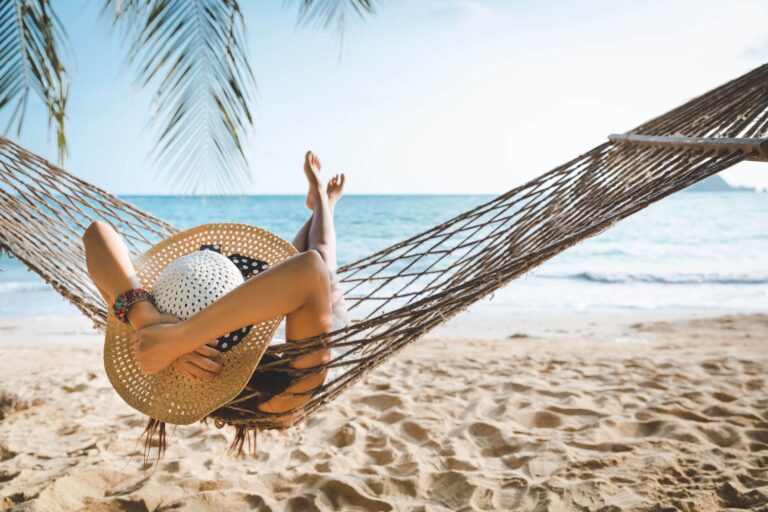 Eine Frau mit Sonnenhut entspannt in einer Hängematte an einem Sandstrand.