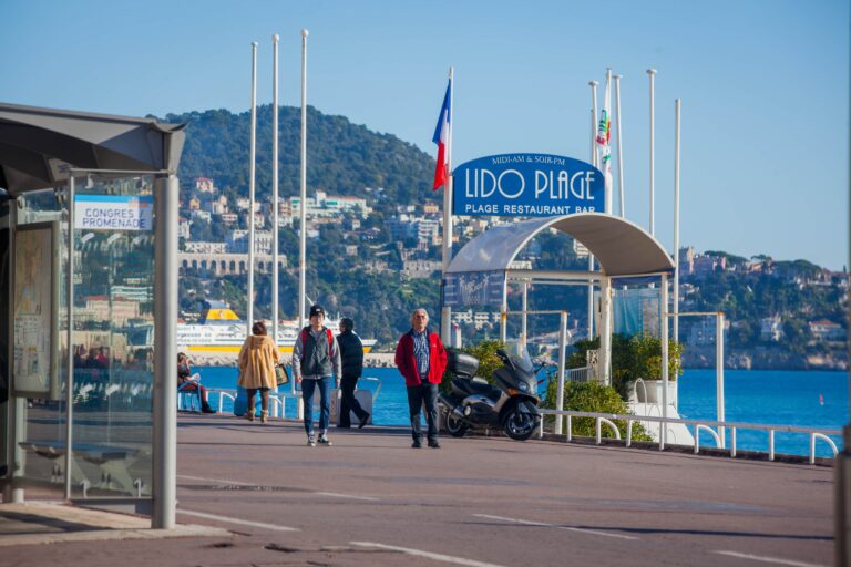 Fußgänger auf der Promenade des Anglais in Nizza neben dem Eingangsschild zum Lido Plage.