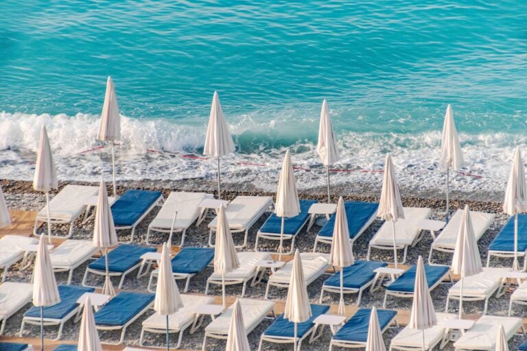 Weiße und blaue Sonnenliegen mit Sonnenschirmen vor türkisfarbenem Meerwasser.