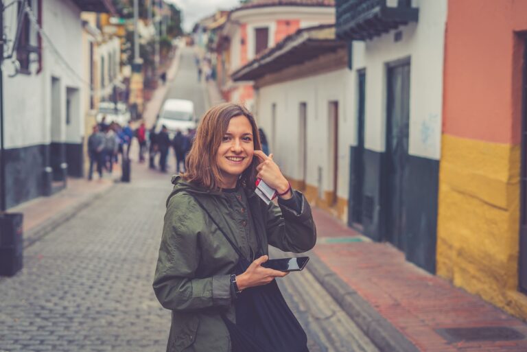 Lächelnde Touristin mit Smartphone in einer bunten, historischen Gasse in Bogotá.