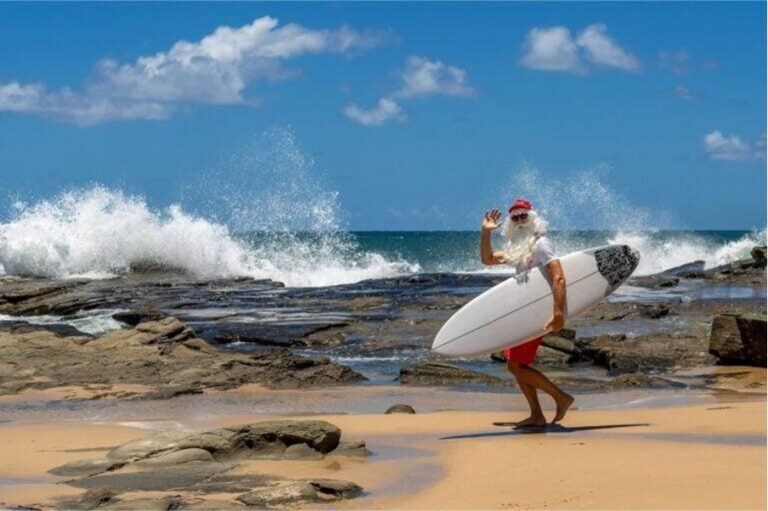 Ein Mann im Weihnachtsmann-Kostüm mit Surfbrett winkt am Strand vor brandenden Wellen