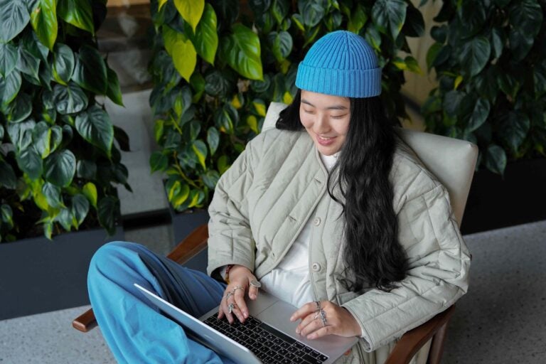 Eine junge Frau mit blauer Mütze sitzt in einem Sessel und arbeitet am Laptop.