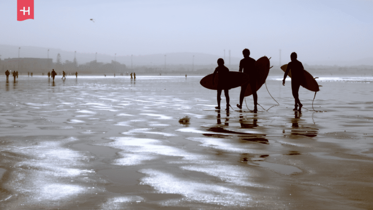 Drei Surfer laufen als Silhouetten mit ihren Surfbrettern über einen weiten Sandstrand.