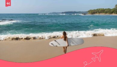 Eine Frau von hinten trägt ein Surfbrett am Sandstrand. Passend zum Thema: Sri Lankas schönste Strände.
