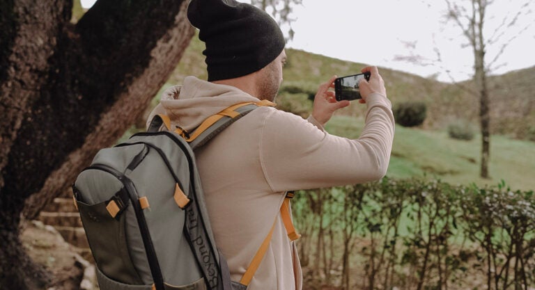 Hombre joven filma con su móvil un paisaje natural. Se ven montañas verdes y el tronco de un árbol. Conexión cuando viajas. 