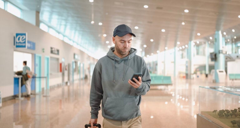 Hombre joven en el aeropuerto mira su celular para resolver algunas necesidades gracias a su conexión a internet.