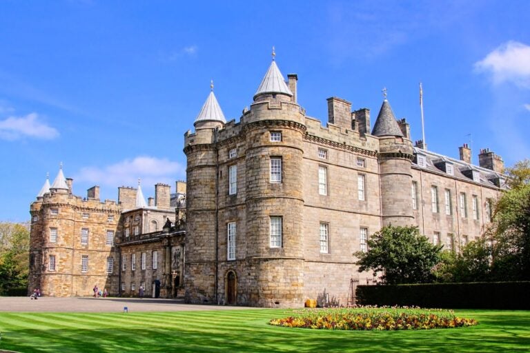 Fachada del Palacio de Holyroodhouse en Edimburgo con jardines verdes y cielo azul despejado.