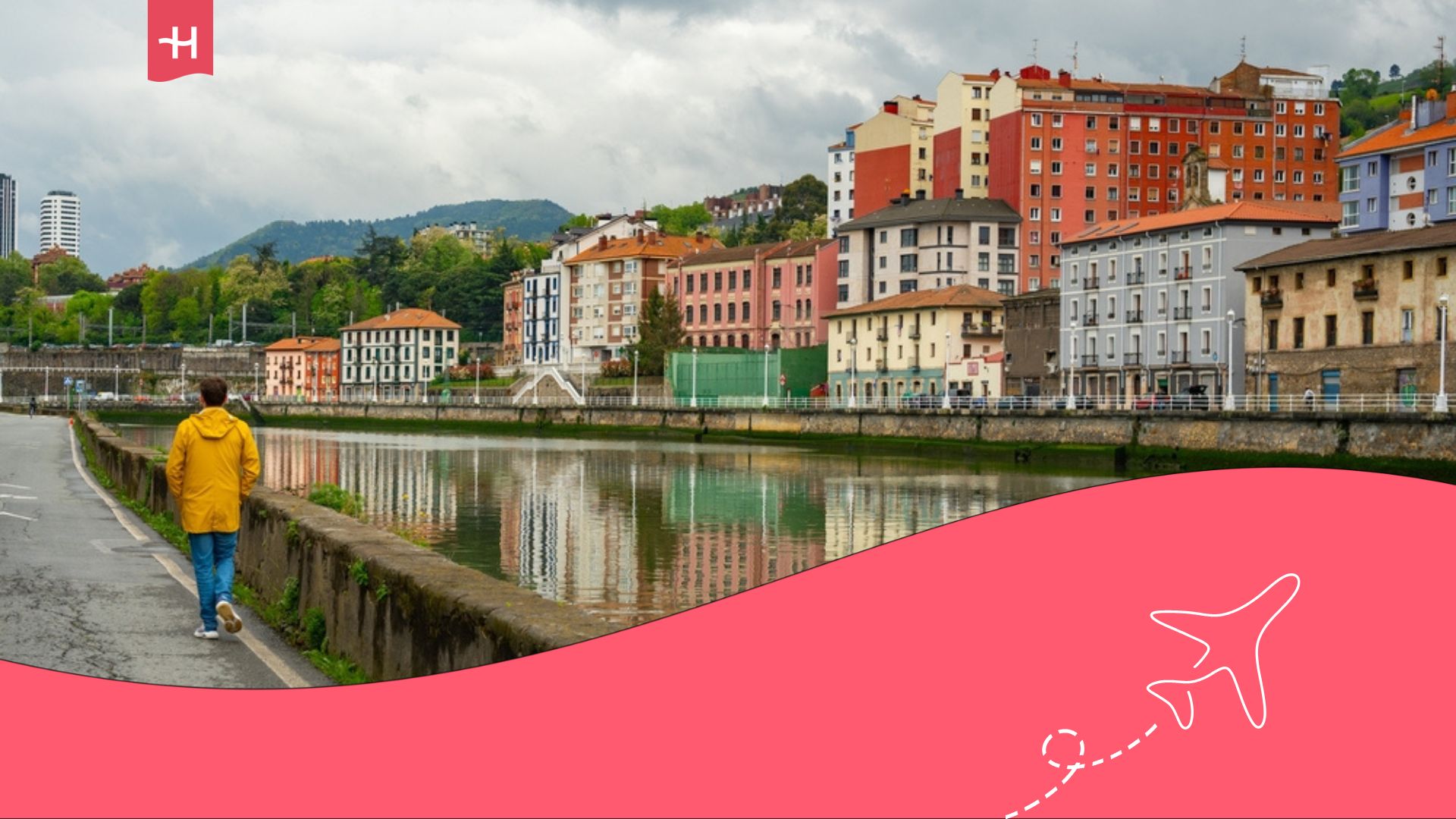 Hombre con chaqueta amarilla caminando cerca del río en Bilbao con casas al fondo.