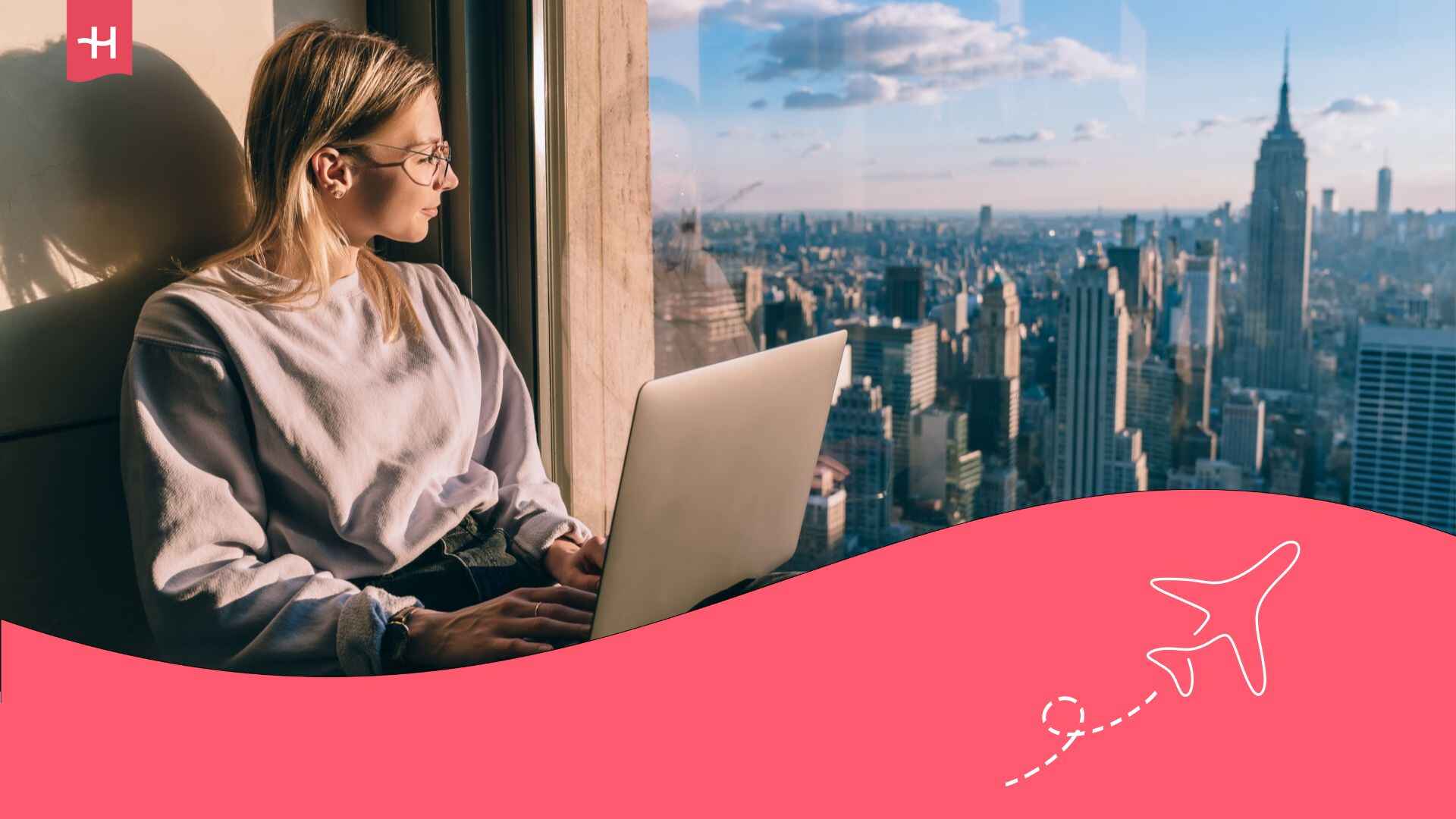 Une jeune femme avec un laptop assise près d'une fenêtre et observant la vue sur les gratte-ciels de New York