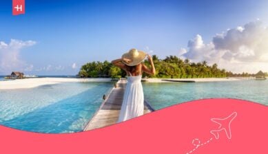 Vista da dietro di una donna elegante in abito bianco e cappello che cammina lungo un molo verso un’isola tropicale alle Maldive