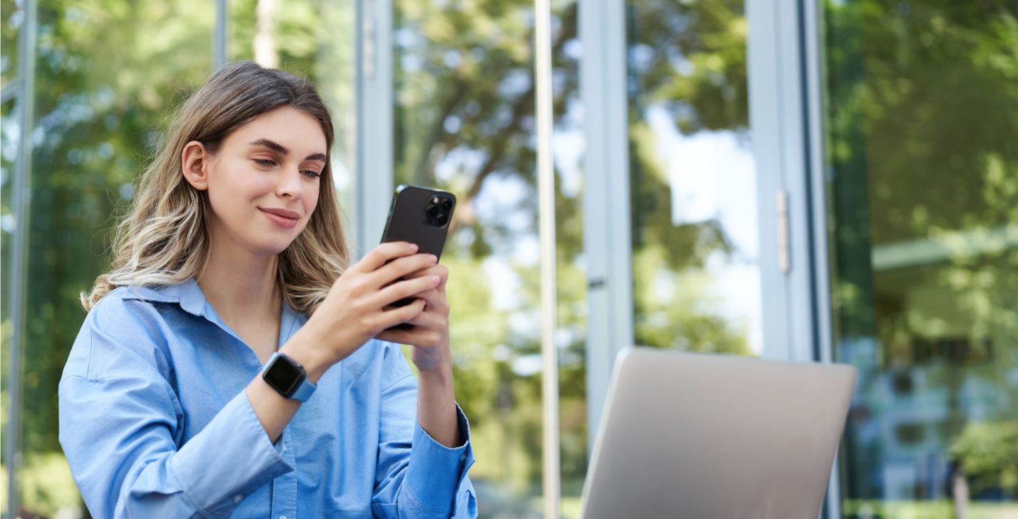 Donna che consulta il suo cellulare con il portatile davanti a sé.